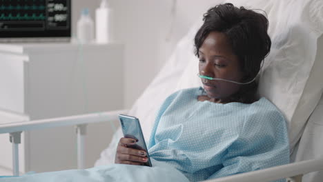 A-young-woman-writes-a-message-on-her-phone-while-lying-in-a-hospital-ward.-An-African-girl-is-lying-in-a-ward-connected-to-ECG-and-oxygen-devices-in-a-mask-and-writes-messages-to-relatives
